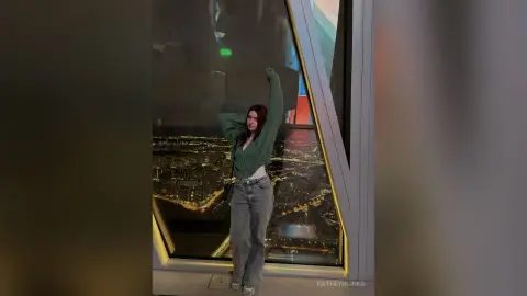 Young woman in a green sweater and jeans posing by a skyscraper window overlooking a glowing city at night.