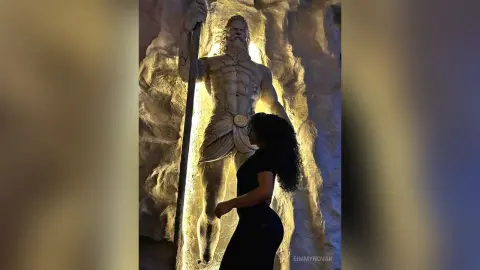 Woman in black outfit standing before a massive, illuminated mythological statue in a dark cave setting.