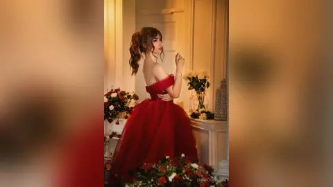 Elegant woman in a red evening gown posing in a romantic floral setting with warm lighting.