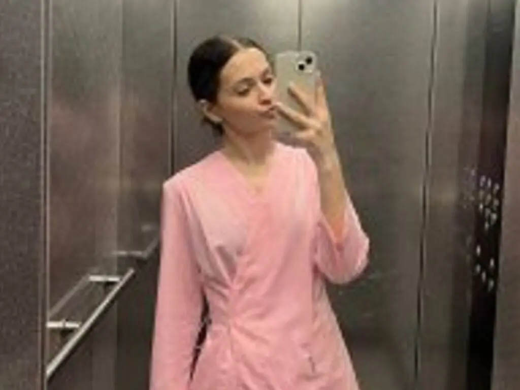Young woman in pink medical scrubs taking a mirror selfie in a modern elevator.