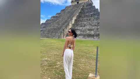 Woman in white trousers and bikini top standing before the ancient Chichen Itza pyramid under a blue sky.