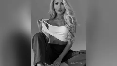 Blonde woman in a white ribbed tank top and black trousers posing in a moody black and white portrait.