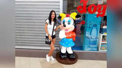 Smiling woman in a white t-shirt and black shorts standing next to a large Minnie Mouse statue outside a shop.