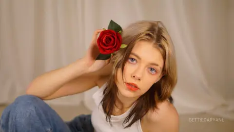 Young woman with a red rose behind her ear wearing a white tank top in a soft studio setting.