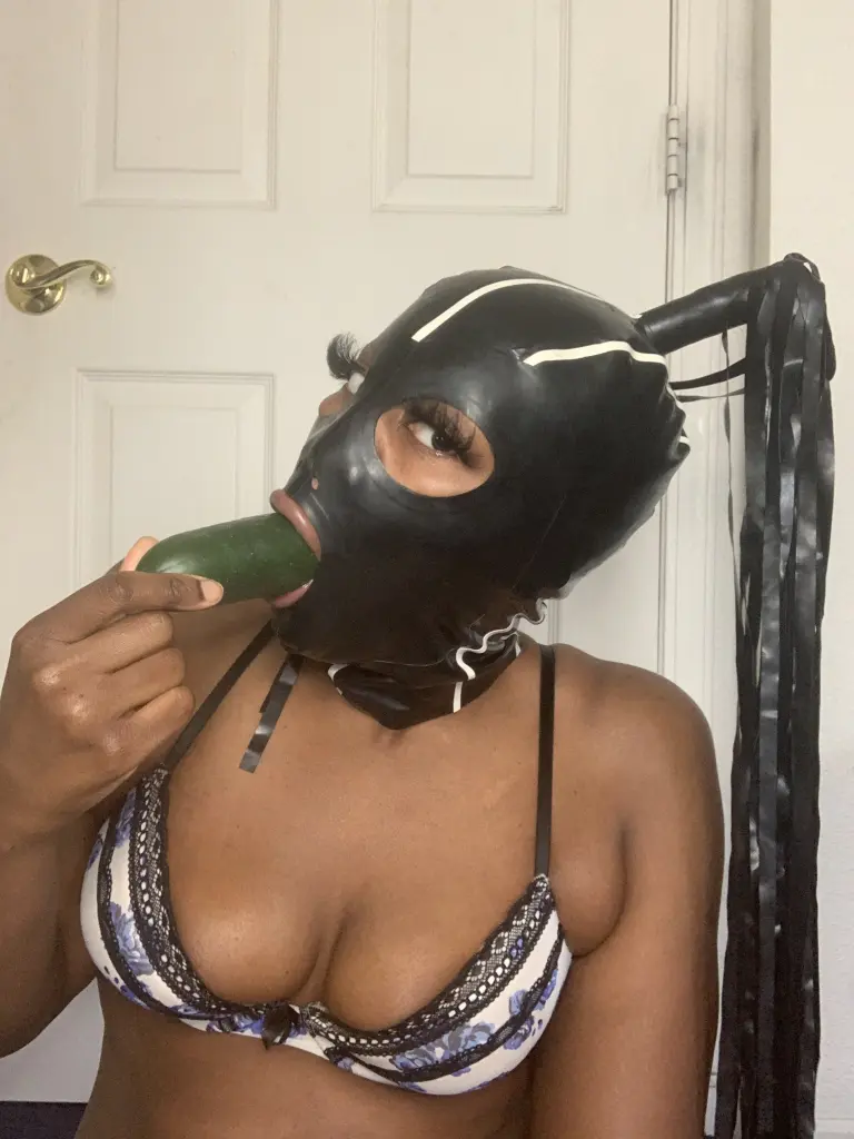 HeadUsa wears a black shiny mask and a black lace top while posing against a plain wall.