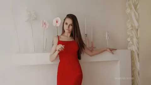 IngridBeckton poses in a red dress with her long brown hair down, leaning against a white mantelpiece in a bright room.