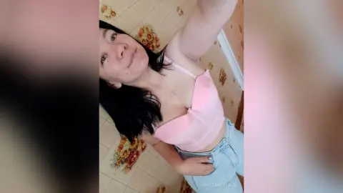 Smiling young woman in a pink camisole and light denim jeans posing in a room with floral wallpaper.