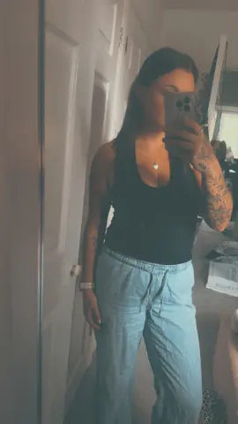 TattedBrat26 takes a mirror selfie wearing a black tank top and light blue sweatpants, showing off her arm tattoos.