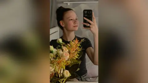 Young woman in a black t-shirt taking a moody mirror selfie next to a bouquet of white and peach flowers.