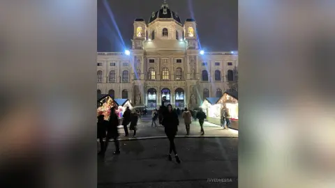 People walking past festive Christmas market stalls in front of a grand illuminated European palace at night.