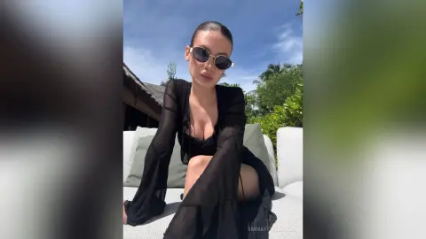 Stylish woman in a black sheer long-sleeve top and sunglasses relaxing on a white outdoor sofa under a sunny sky.