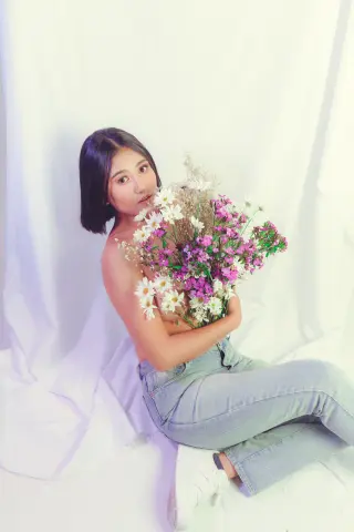 Young woman in light denim jeans holding a bouquet of purple and white wildflowers in a bright studio setting.