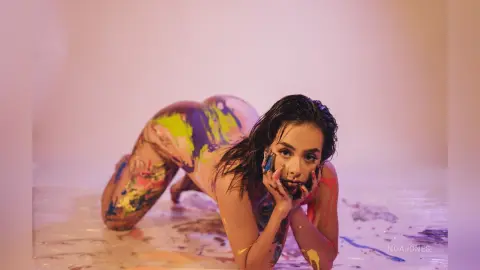 Artistic woman covered in colorful paint posing on a studio floor in a dreamy, creative mood.