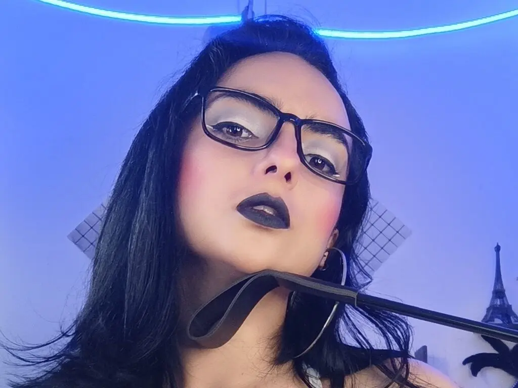 Woman with dark hair and black lipstick wearing glasses in moody blue lighting.