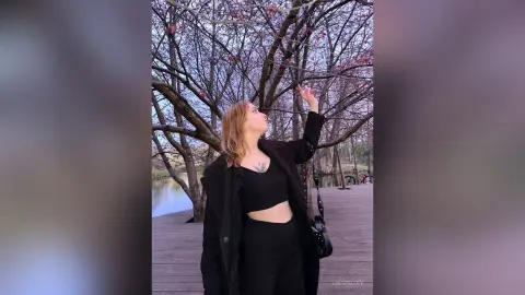 OksiKoxy stands outdoors on a wooden walkway, wearing a black crop top and coat while looking up at the branches of a flowering tree.