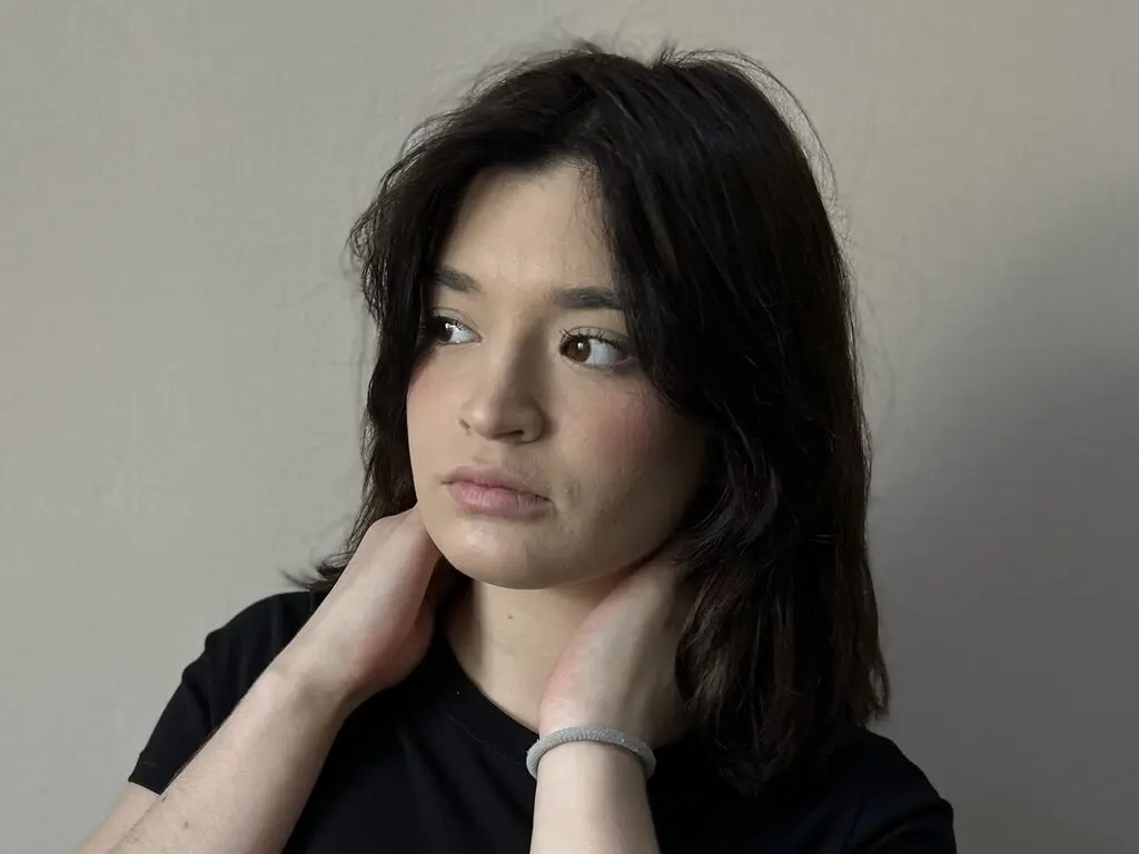 Young woman with wavy dark hair wearing a black t-shirt looking thoughtfully against a neutral wall.