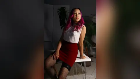 Young woman with pink hair in a white sleeveless top and red skirt sitting in a moody, dimly lit room.