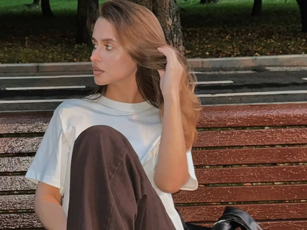 LeslieDorian sits on a wooden bench outdoors, wearing a white t-shirt and dark pants while running her hand through her long brown hair.