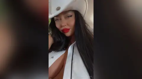 Glamorous woman with long dark hair and red lipstick wearing a white cowboy hat and outfit.