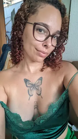 dominamoon — Smiling woman with curly hair and butterfly chest tattoo wearing a green lace camisole in a bright room.