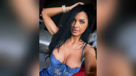 Beautiful woman with long dark hair wearing blue lace lingerie in a soft, intimate bedroom setting.