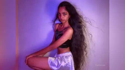 Young woman with long wavy hair wearing a black satin camisole and white shorts in moody purple lighting.