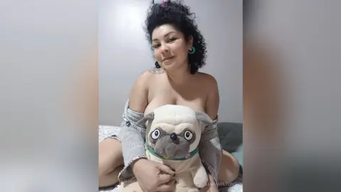 Smiling woman with curly hair sitting in a cozy room holding a large pug plushie toy.