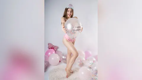 Ada — Cheerful young woman in pink lingerie and tiara holding a Happy Birthday balloon amidst pink party decorations.