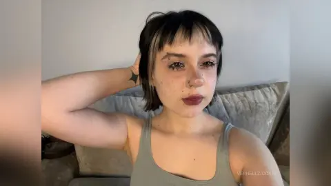 VernellZiccardi has short dark hair and wears a grey tank top while posing with one hand behind her head against a grey couch.