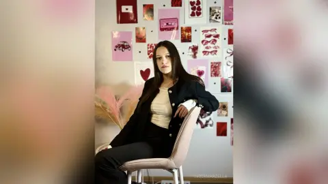 IngerMarzella sits in a chair wearing a black cardigan over a white top, posing in front of a wall decorated with heart and pink-themed prints.