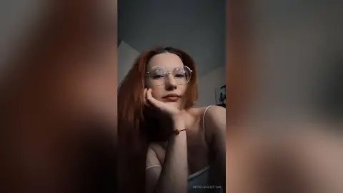 Redheaded woman with glasses in a white camisole posing thoughtfully in a dimly lit room.