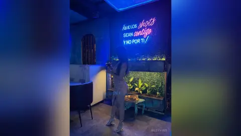 Woman in a stylish outfit standing by a neon sign and greenery in a moody, blue-lit lounge bar.