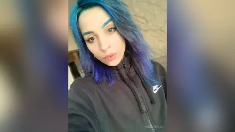 Young woman with blue hair wearing a black Nike sweatshirt in a casual indoor setting.