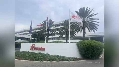 Modern white hotel building with palm trees and flags under a cloudy sky.