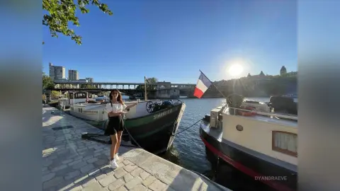 DyandraEve stands on a stone walkway next to a boat in a river under a bright sun.