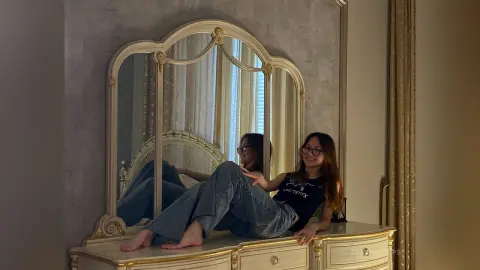 Smiling woman in a black graphic t-shirt and blue jeans relaxing on a vintage white vanity in a cozy room.