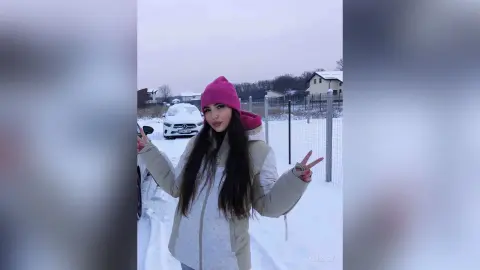 Young woman in a beige winter jacket and pink beanie making peace signs in a snowy outdoor setting.