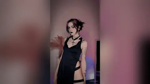 LorenLoiss poses in a black cut-out bodysuit and black choker, showing her tattoos and dark hair against a plain wall.