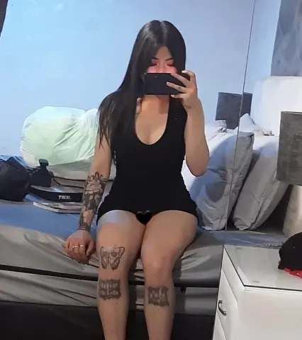 Tattooed woman in a black bodysuit sitting on a bed in a cozy bedroom taking a mirror selfie.