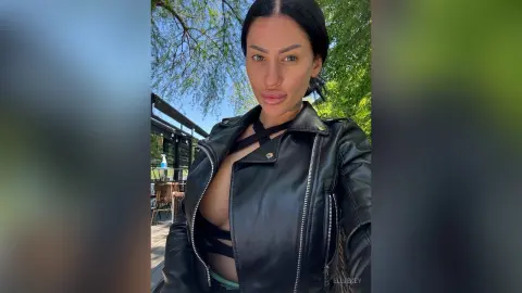 Confident woman in a black leather biker jacket posing outdoors against a lush green leafy background.