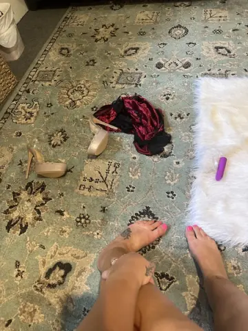 LexiLotusUS lies on a patterned rug with her legs bent, wearing light tan high heels and showing her bare feet and legs.