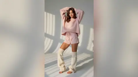 Young woman in pink loungewear and white leg warmers posing in a bright, minimalist studio setting.