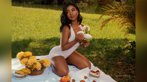 Beautiful woman in a white bodysuit sitting on a picnic blanket with flowers in a sunny garden setting.