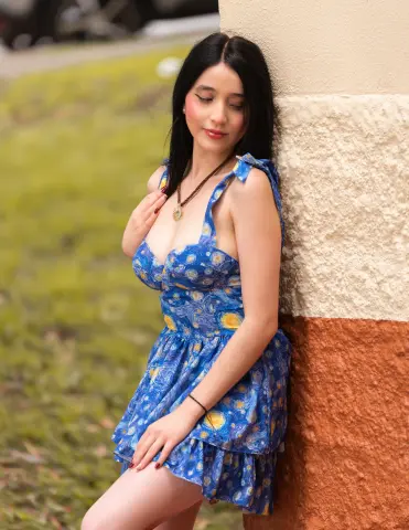 Young woman in a blue floral sundress leaning against a textured wall in a sunny outdoor garden setting.