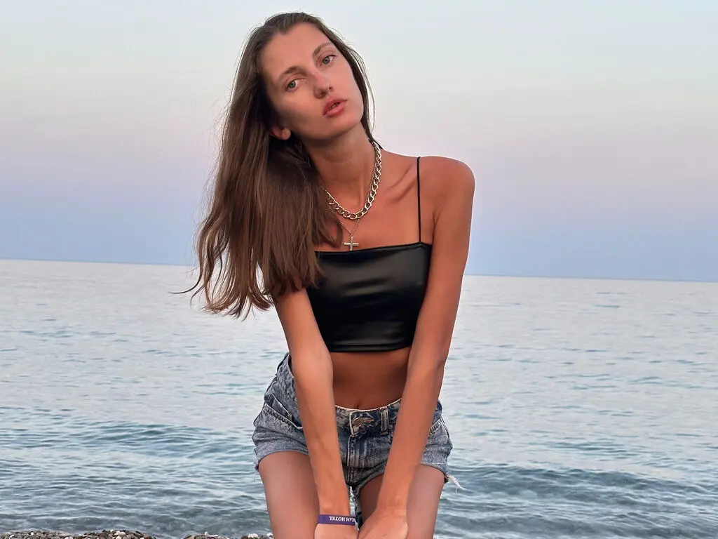 Young woman in black crop top and denim shorts relaxing by the ocean at sunset.