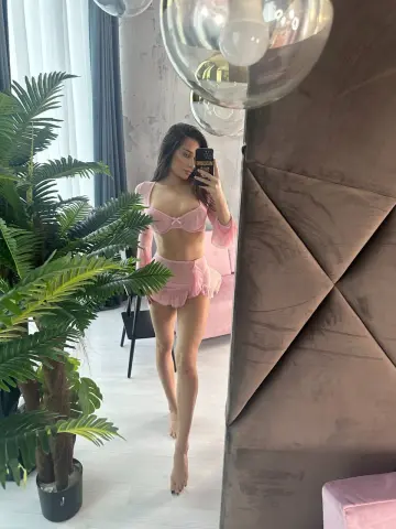 Young woman in pink ruffled lingerie set posing in a modern bedroom with a large indoor plant.