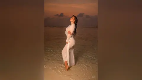 Elie poses on a beach at sunset wearing a long white dress that hugs her curves, standing in shallow water.