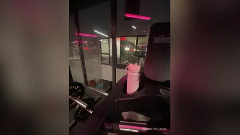 ChristineDavis poses in a gym setting, holding a pink water bottle near a weight rack and treadmill.