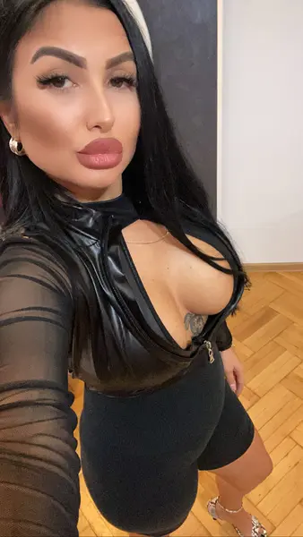 mistress_meryem poses naked on a silver sofa, wearing black sheer mesh stockings and a black bodysuit that shows her cleavage and bare skin.