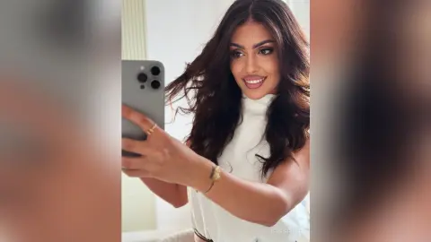 Smiling brunette woman in a white sleeveless turtleneck taking a mirror selfie in a bright indoor setting.